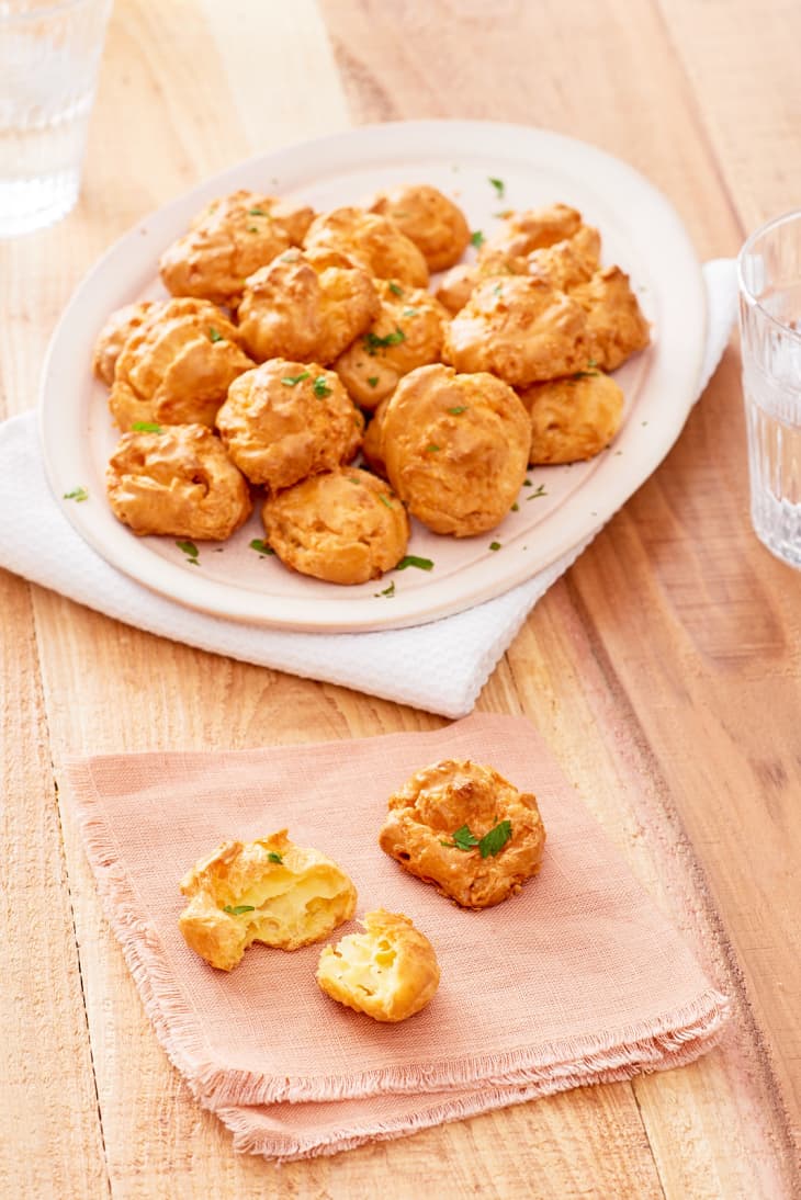 Freshly baked cheese gougères are piled up on a white plate, while a piece on a peach-colored napkin is torn to pieces