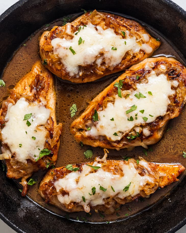 Chicken breasts topped with melted cheese and caramelized onions in a skillet, garnished with fresh herbs.