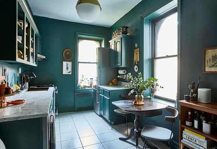 Teal kitchen with marble countertops, vintage decor, wooden table, chairs, and plants by the window.