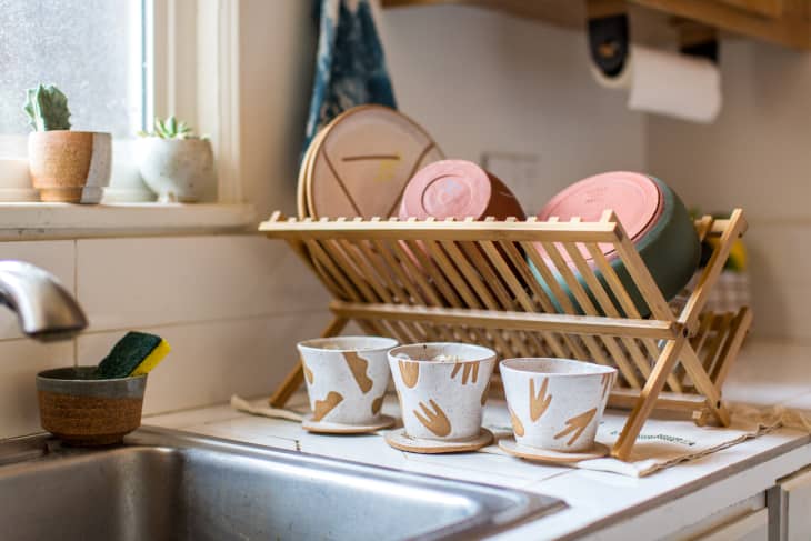 Dish rack with pink and green bowls, cups with handprint designs, and potted plants by a kitchen sink.