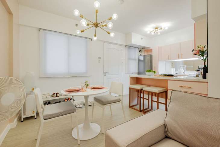 Bright dining area with a round table, four chairs, and a modern chandelier, leading to a kitchen with pink cabinets.