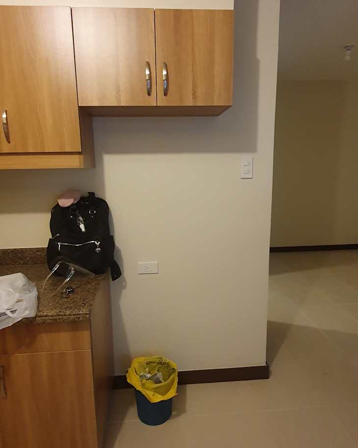 Kitchen corner with wooden cabinets, a black backpack, and a blue trash can with a yellow bag.