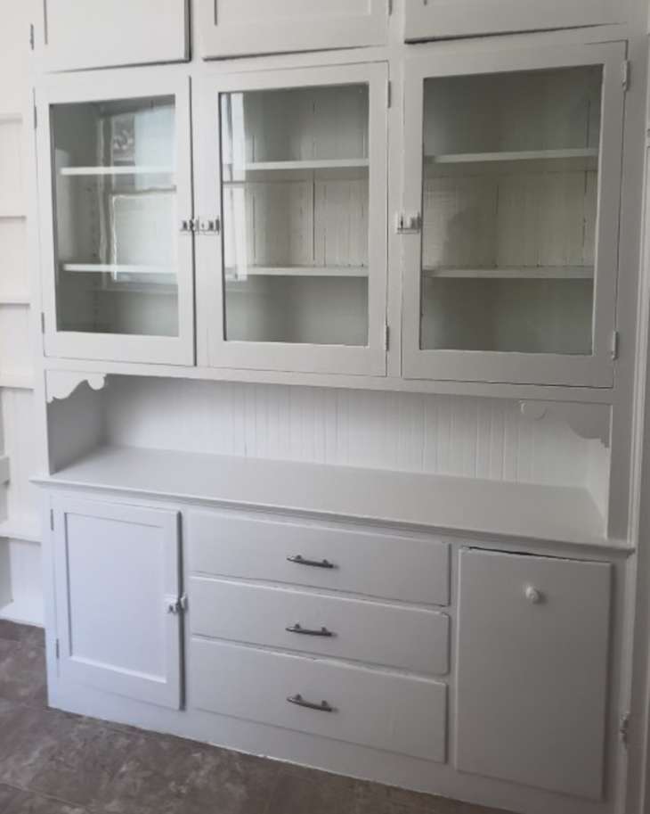 White wooden cabinet with glass doors, open shelves, and three drawers, featuring a smooth countertop.
