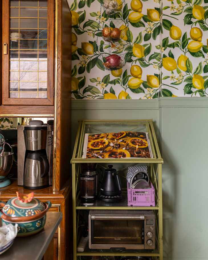 Kitchen corner featuring a green shelf with a cookbook, coffee maker, kettle, and an oven, surrounded by lemon-patterned wallpaper.