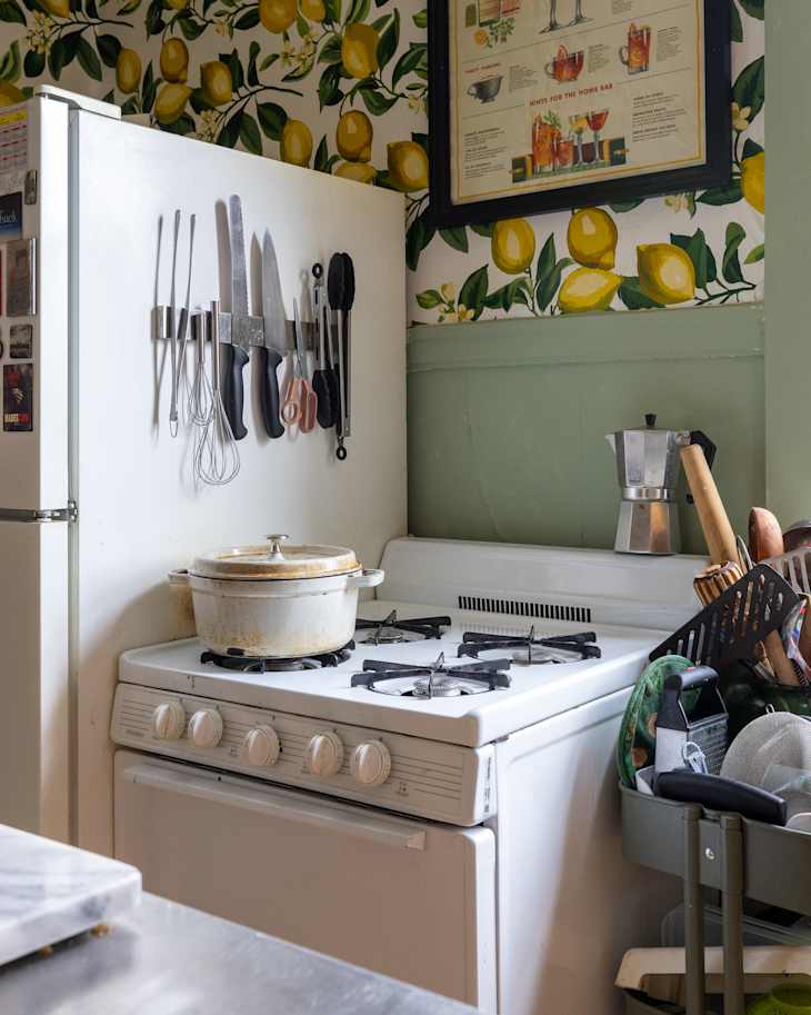 Kitchen corner featuring a white gas stove with a pot, hanging utensils, and a green cart filled with cooking tools.