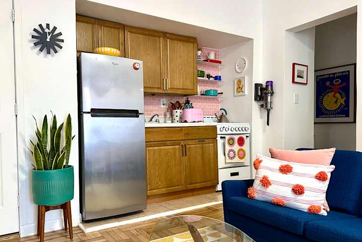 Modern kitchen with stainless steel fridge, wooden cabinets, pink tile backsplash, and colorful decor. Blue sofa with decorative pillows.