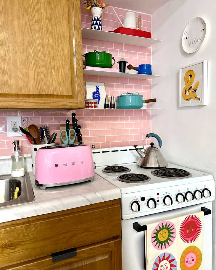 Bright kitchen featuring a pink SMEG toaster, white stove, green and blue cookware on shelves, and colorful dish towels.