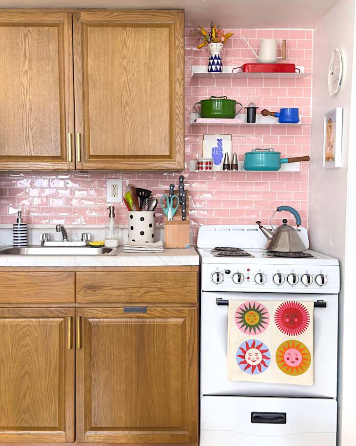 Bright kitchen corner with wooden cabinets, pink tiled backsplash, colorful cookware on shelves, and a white stove with a decorative towel.