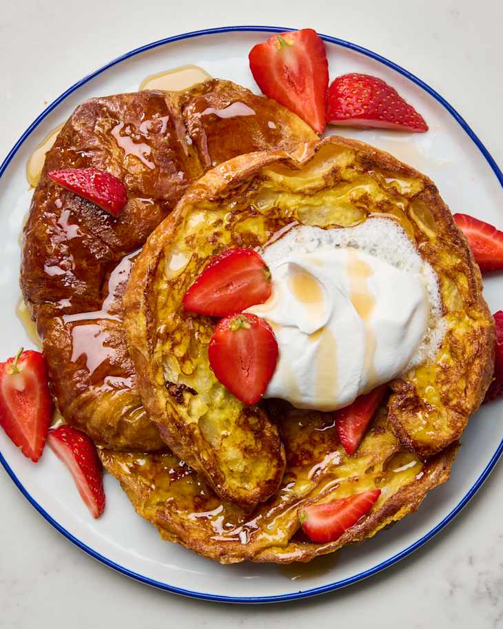 Golden croissant French toast topped with whipped cream, fresh strawberries, and syrup on a white plate.
