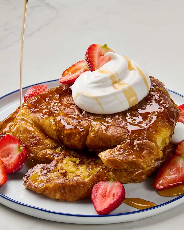 Golden croissant French toast topped with whipped cream, fresh strawberries, and syrup on a white plate.