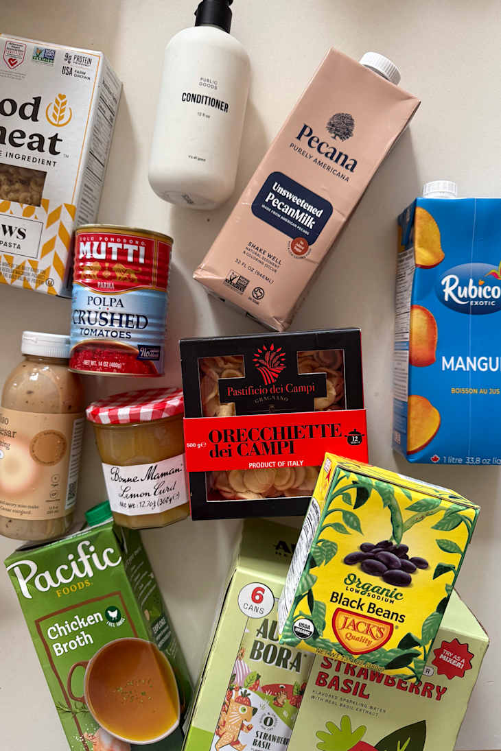 Various food products including pasta, crushed tomatoes, pecan milk, chicken broth, and lemon curd on a countertop.
