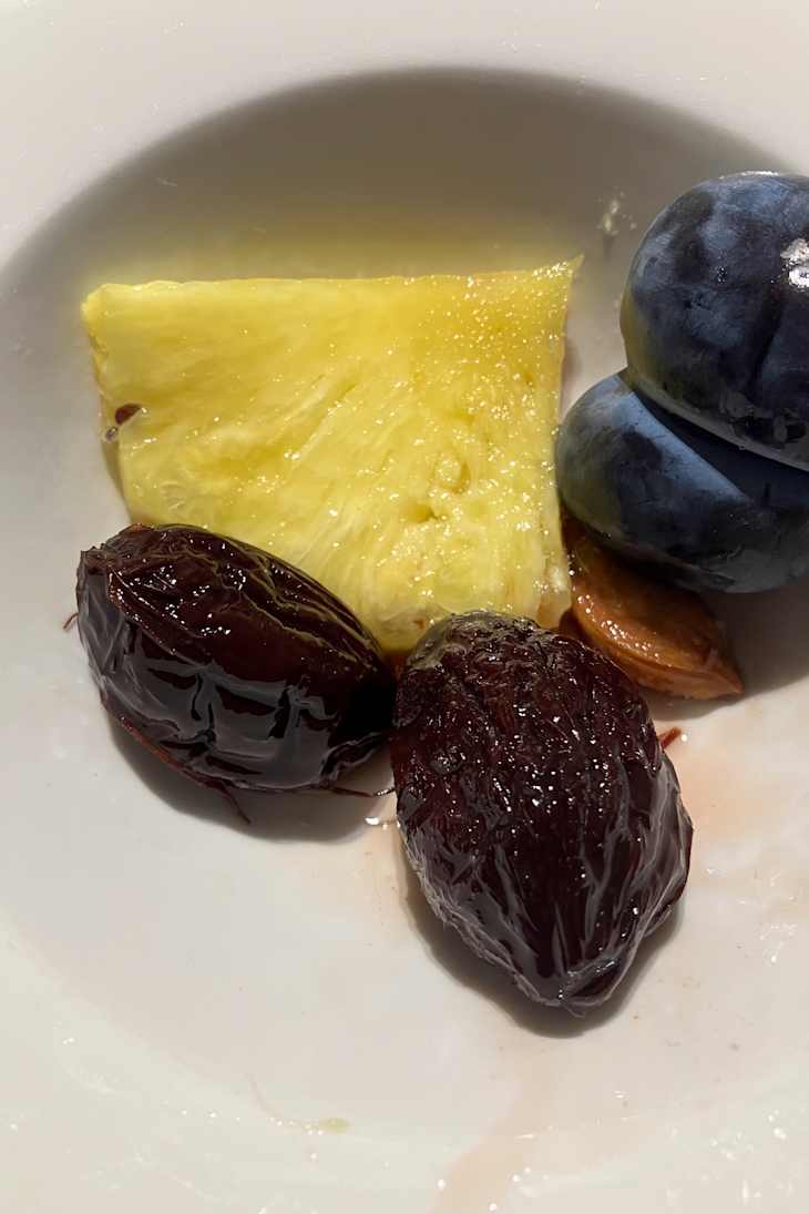 A slice of pineapple, two dark dates, and three plump plums on a plate.
