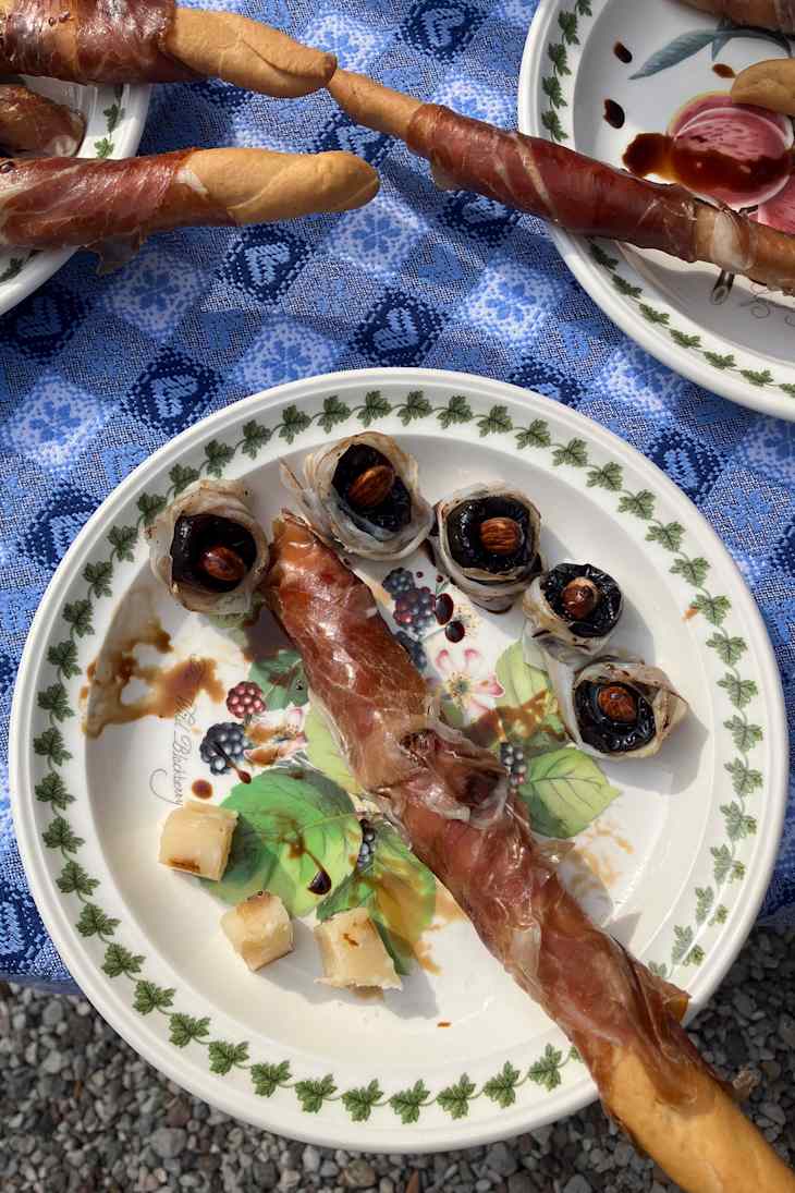 A plate with a prosciutto-wrapped breadstick, black olives with almonds, and small cheese cubes on a blue patterned tablecloth.