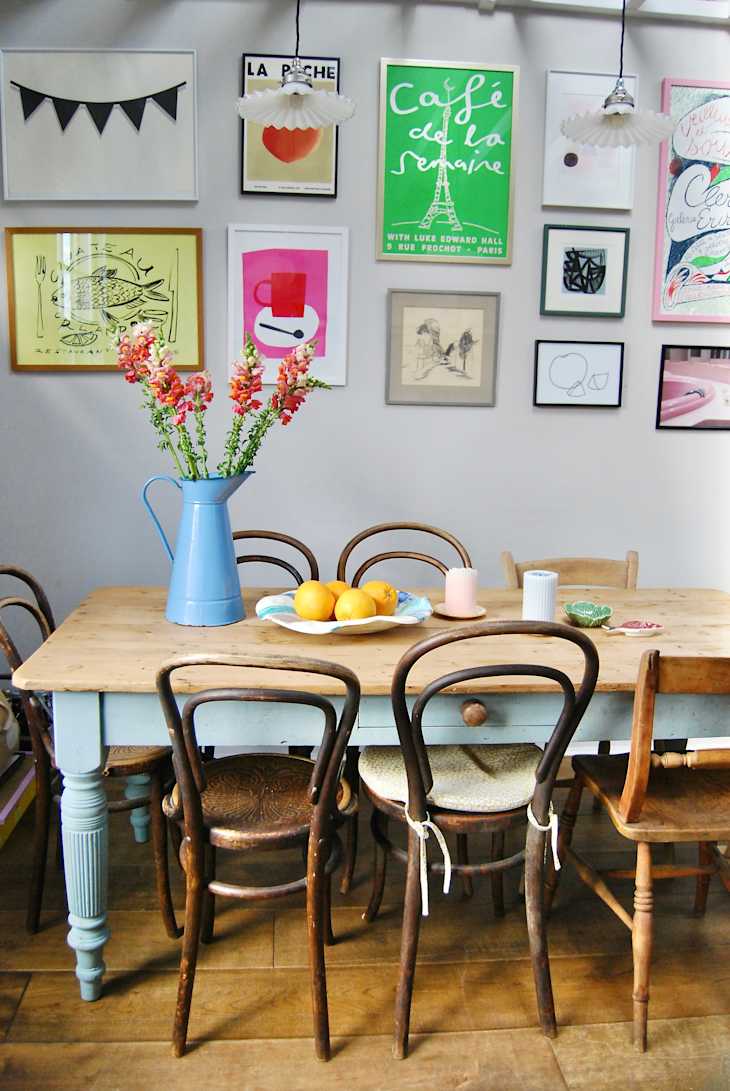 Dining room with wooden table, mismatched chairs, blue vase with flowers, and colorful framed art on the wall.
