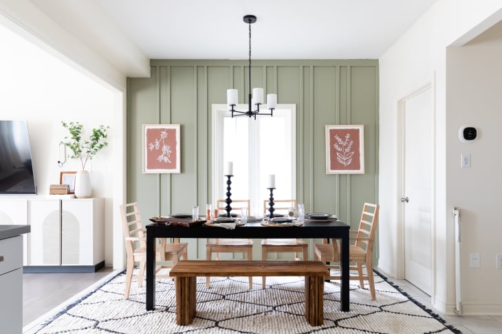 Dining room with black table, wooden chairs, bench, green accent wall, framed botanical prints, and a geometric rug.