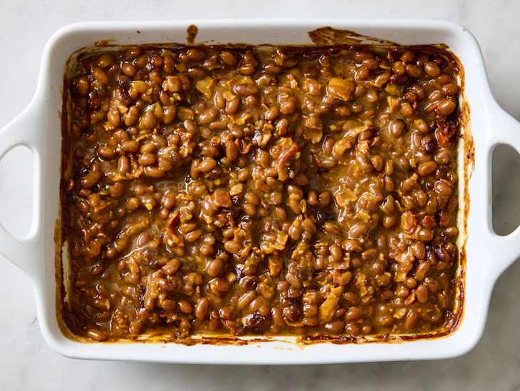 Baked beans in a white casserole dish with a glossy sauce, showing a mix of beans and bits of meat.