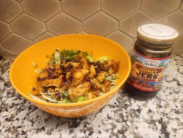Bowl of seasoned chicken with lettuce and red onion beside a jar of Jamaican honey jerk seasoning on a granite countertop.