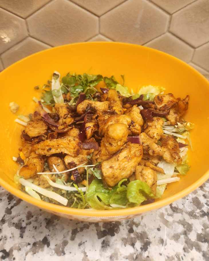 Bowl of mixed salad with grilled chicken, red onions, and various greens on a speckled countertop.