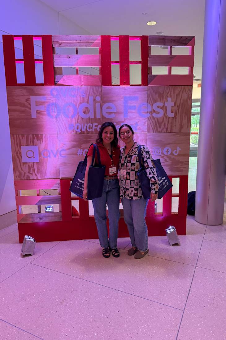 Two women posing together in front of a colorful wooden backdrop with "Foodie Fest" signage, both holding tote bags.
