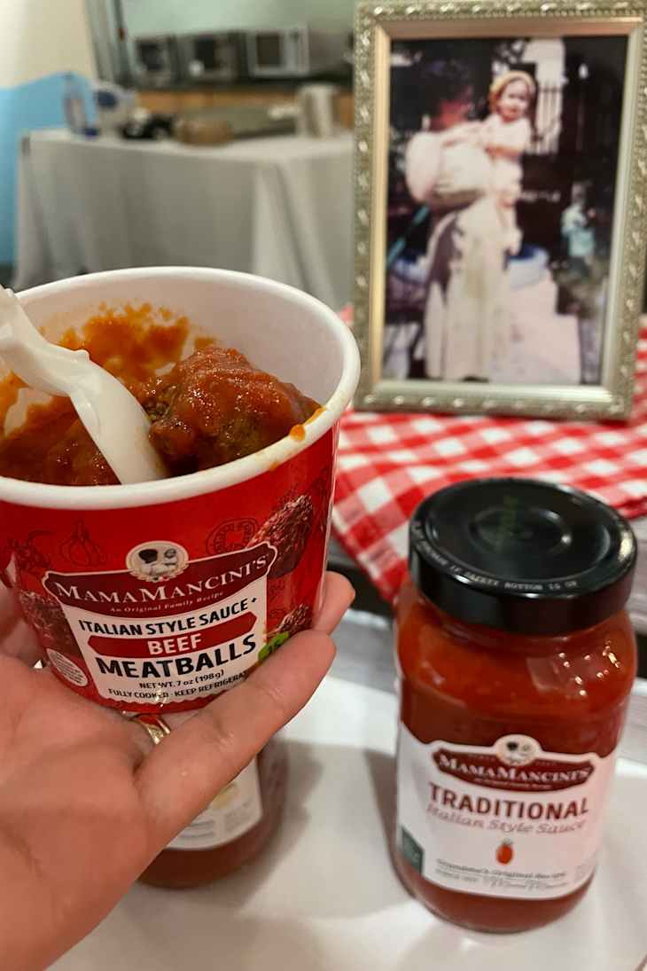 A hand holding a cup of beef meatballs in sauce, with a jar of traditional sauce nearby.