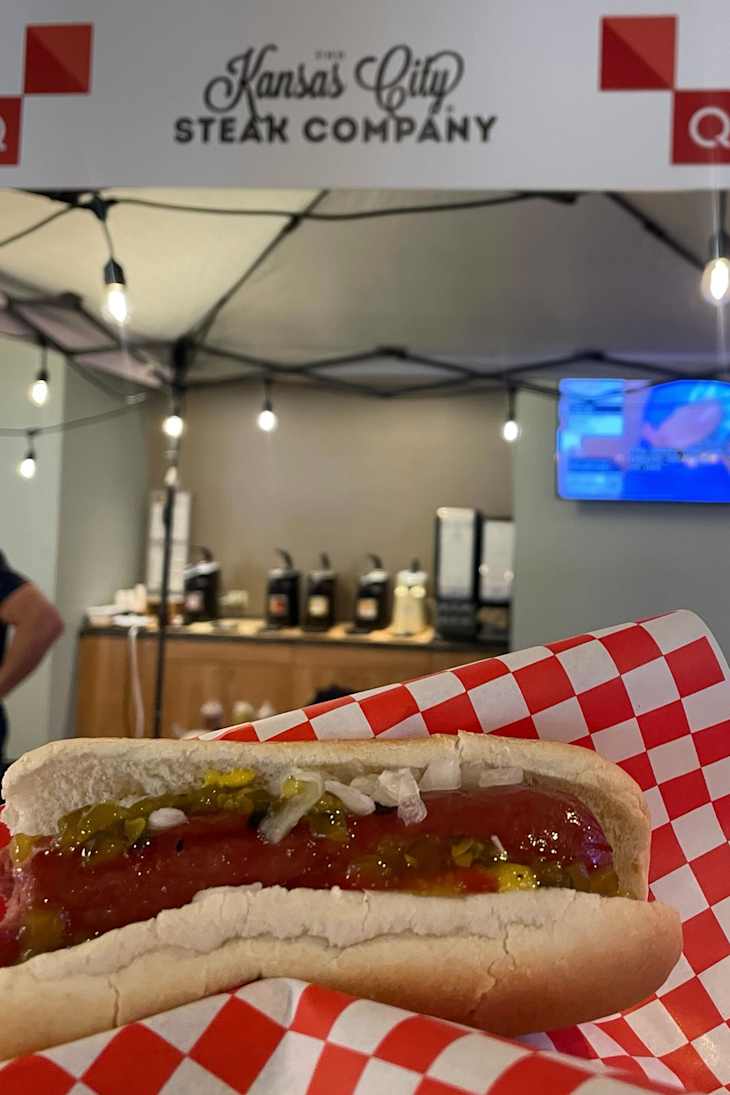Hot dog with ketchup, mustard, and onions in front of the Kansas City Steak Company booth.