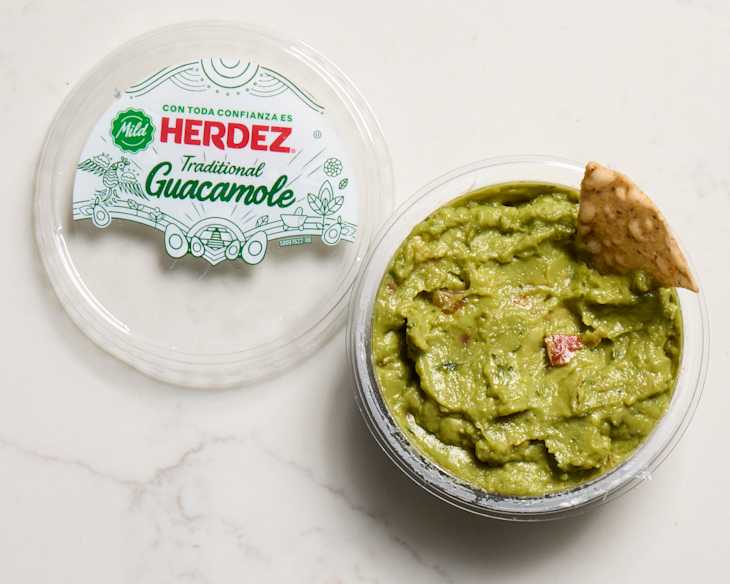 Container of Herdez Traditional Guacamole with a tortilla chip resting on the edge, set on a light surface.