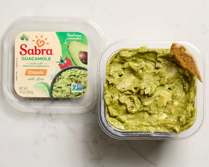 Container of Sabra guacamole with lime, showing creamy green dip and a tortilla chip resting on the edge.