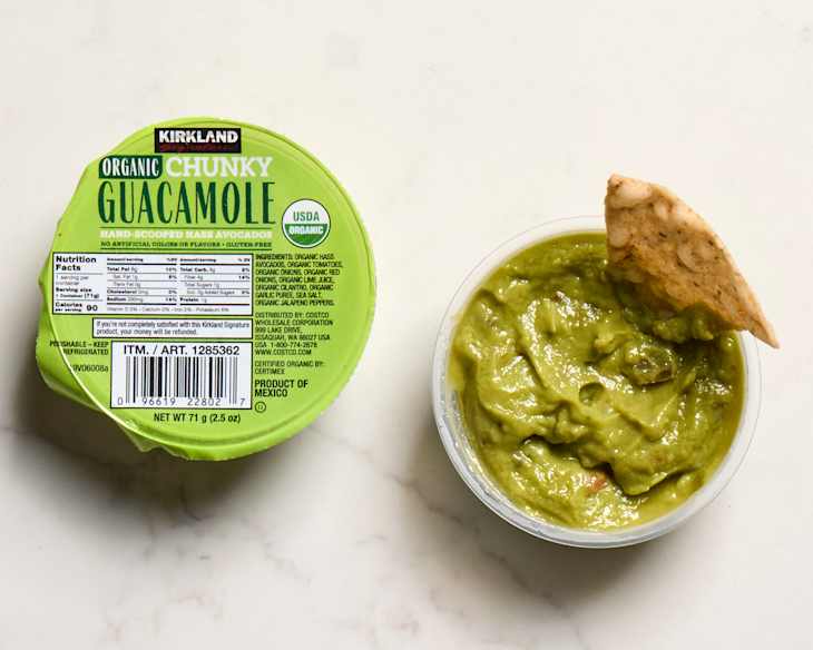 Container of Kirkland organic chunky guacamole with a tortilla chip dipped in, on a light surface.
