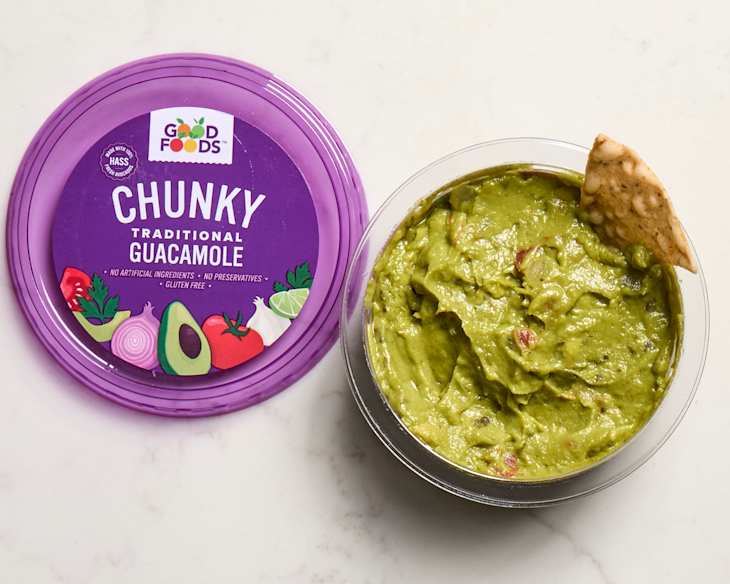 Chunky traditional guacamole by Good Foods in a clear container with a purple lid, accompanied by a tortilla chip.