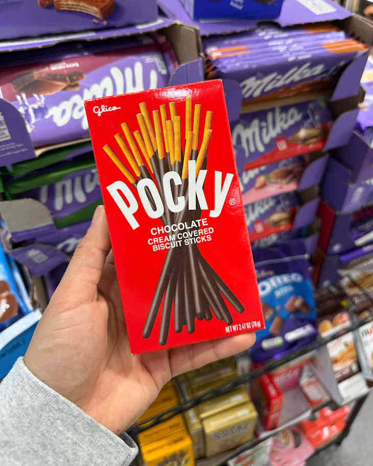 A hand holding a box of Pocky chocolate-covered biscuit sticks, with various snack products in the background.