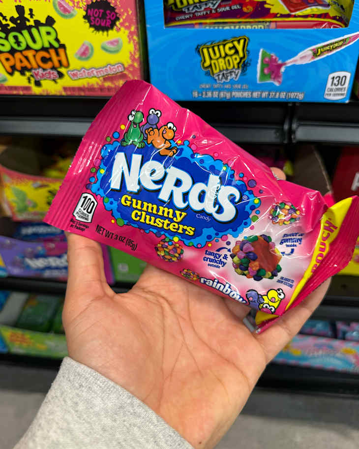 Hand holding a pink package of Nerds Gummy Clusters, featuring colorful candy graphics, in a store aisle.