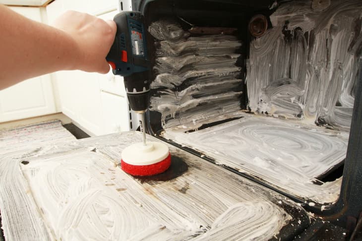 Hand using a power drill with a scrubber attachment to clean an oven interior coated with cleaning paste.