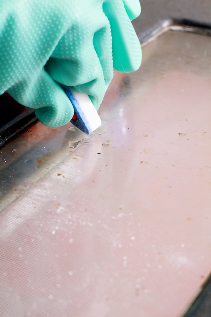Gloved hand scrubbing a dirty oven door with a cleaning sponge.