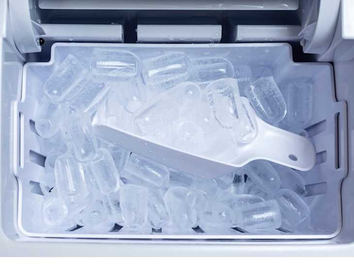 Top view of an ice cube tray full of bullet ice with a white scoop inside a portable automatic ice maker.