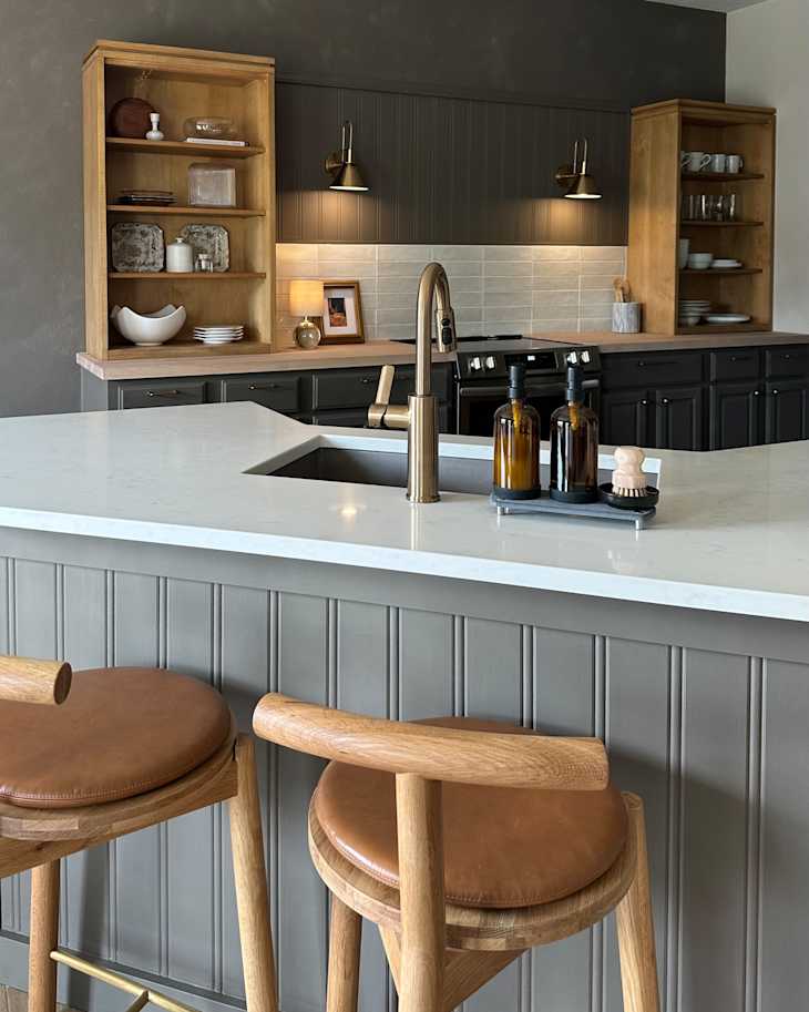 Modern kitchen with gray cabinetry, wooden shelves, a marble countertop, and two wooden bar stools with leather seats.