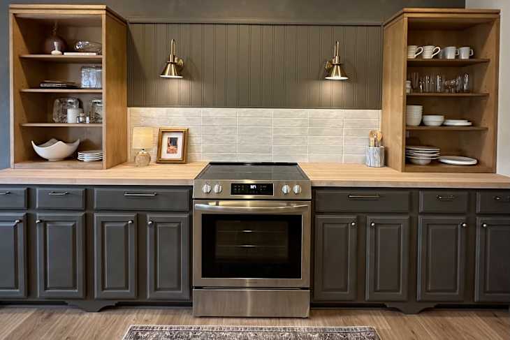 Modern kitchen with dark cabinets, stainless steel oven, wooden countertop, and open shelving displaying dishes and decor.