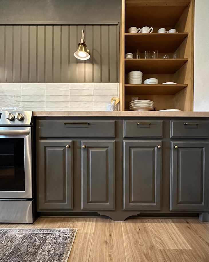 Modern kitchen with gray cabinets, stainless steel stove, wooden shelves displaying white dishware, and a light fixture.