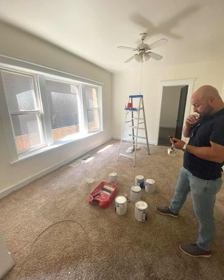 Room undergoing renovation with a man holding keys, paint cans, a roller tray, and a ladder. Bright windows and carpeted floor.