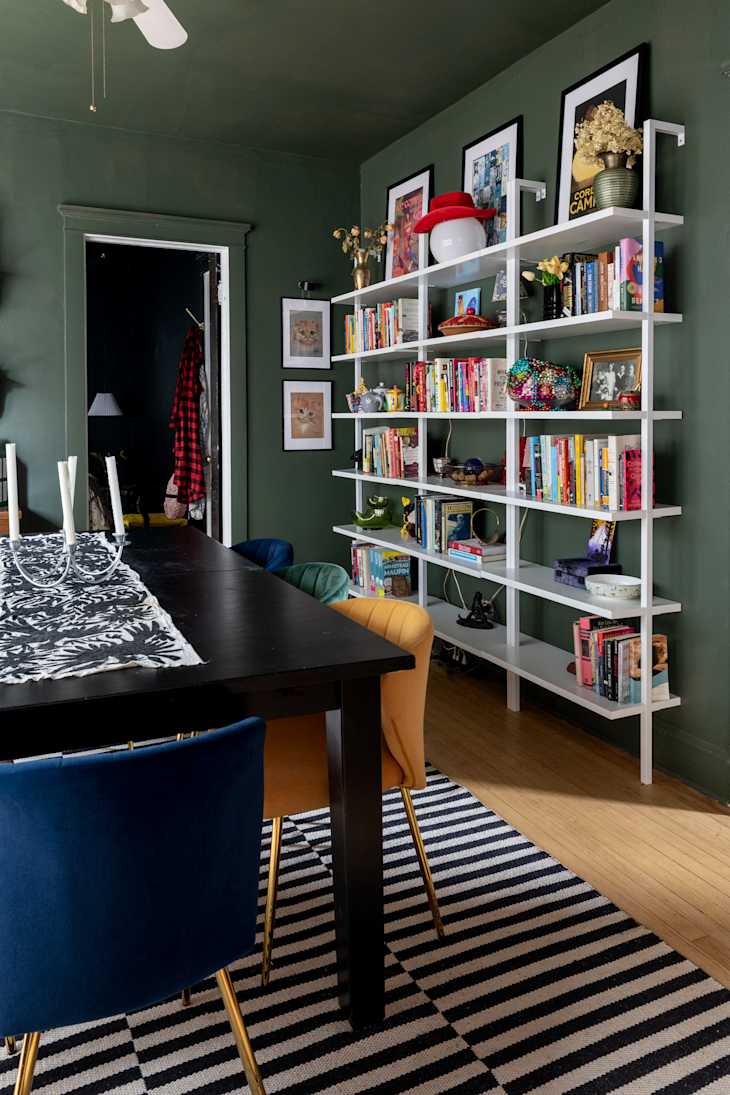 Dining area with a black table, colorful chairs, a striped rug, and a white bookshelf filled with books and decor.