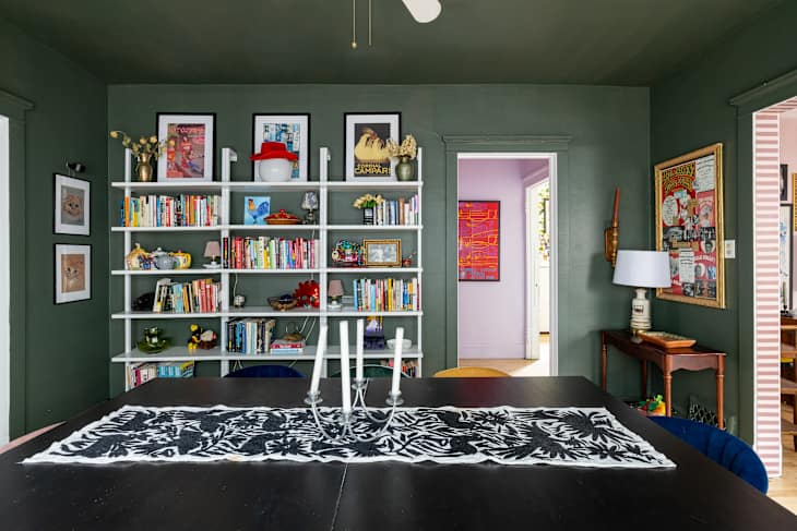 Dining area featuring a dark green wall, a white bookshelf filled with books and decor, and a black table with a patterned runner and candles.