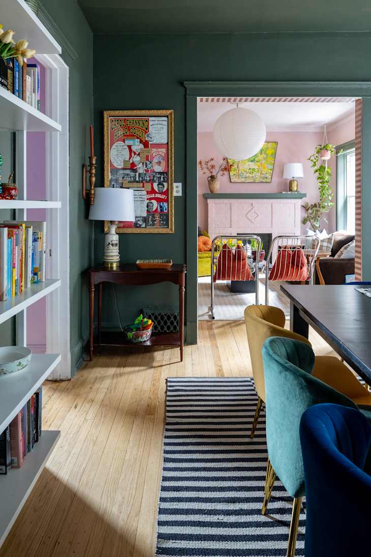 Cozy dining area with colorful velvet chairs, a striped rug, and a view into a pink living room with plants and artwork.