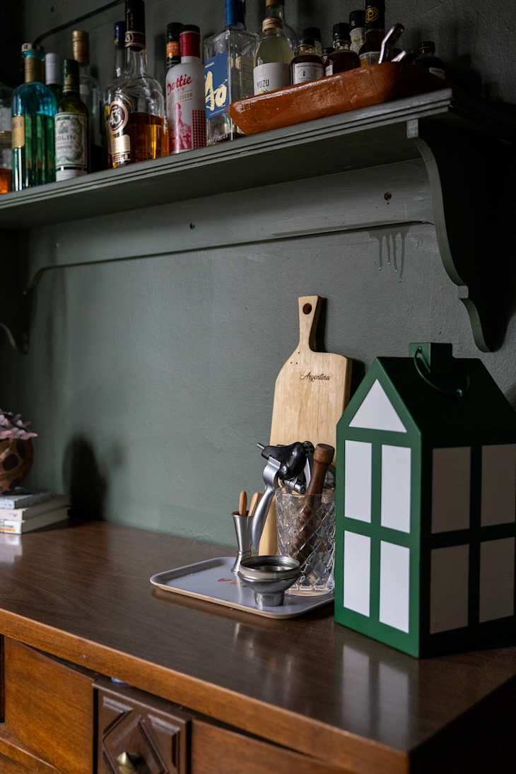 Shelf with various liquor bottles, and a cutting board and bar tools on a wood sideboard in dark dining room.