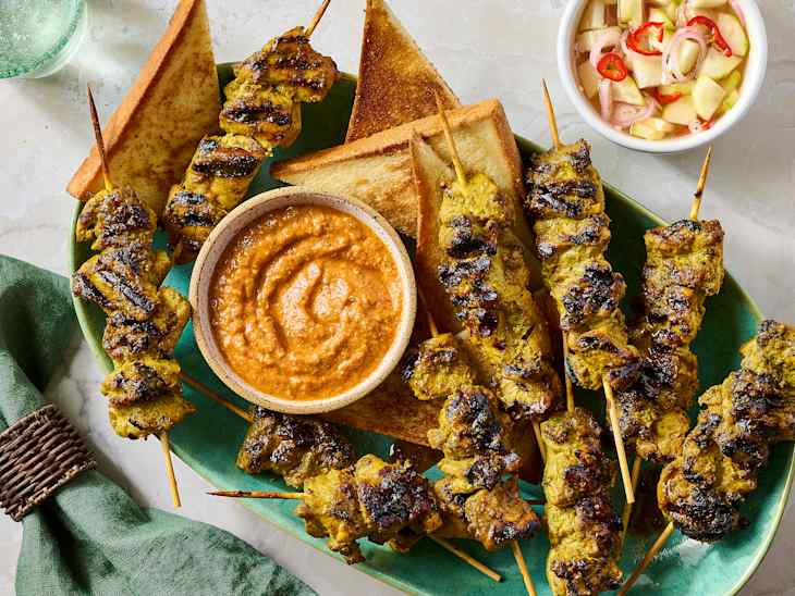 Grilled chicken skewers on a green platter with toasted bread, a bowl of orange sauce, and a side of pickled vegetables.