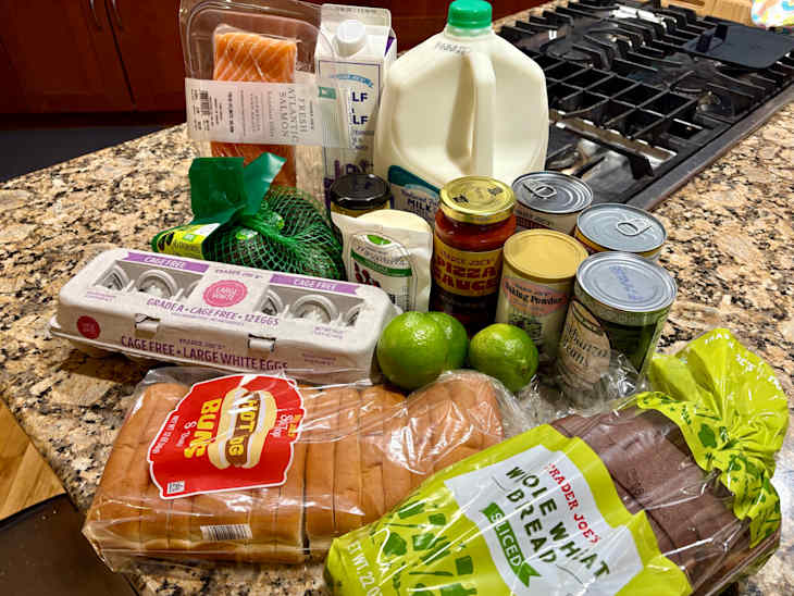 Grocery items on a granite countertop including salmon, eggs, milk, avocados, limes, and various canned goods.