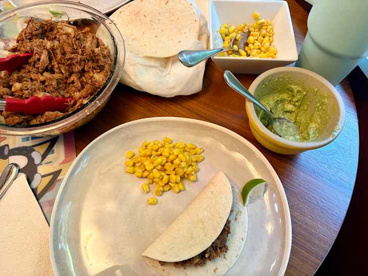 Pulled meat in a bowl with tongs, tortillas, corn, and guacamole on a wooden table.