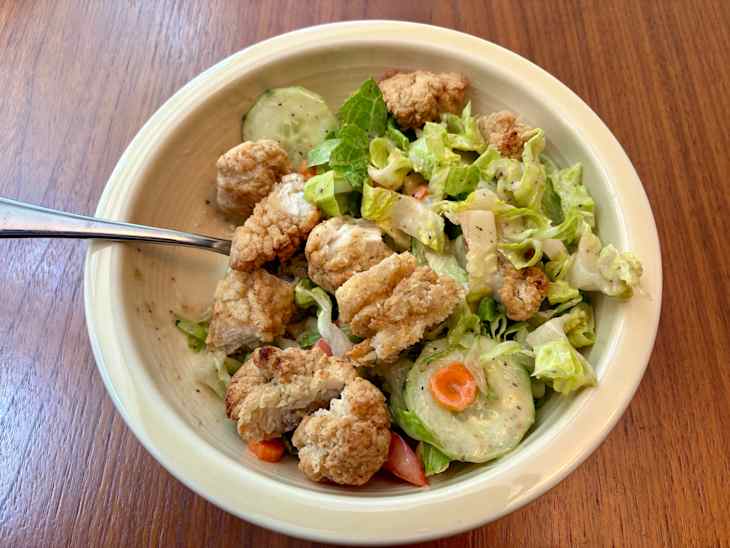 Bowl of salad with crispy fried pieces, lettuce, cucumber slices, and carrots on a wooden table.