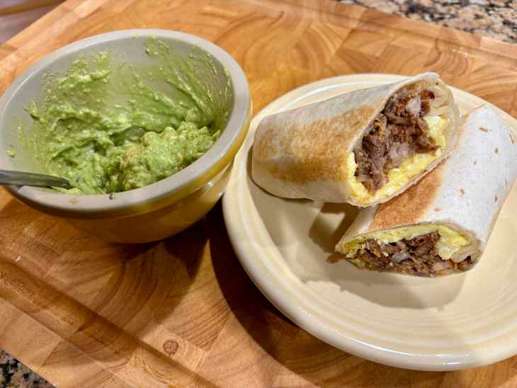 Two breakfast burritos filled with eggs and meat on a plate, alongside a bowl of guacamole.