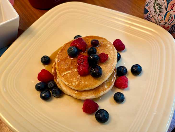 Fluffy pancakes stacked with blueberries and raspberries, drizzled with syrup on a cream-colored plate.