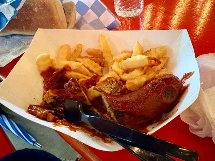 A serving of crinkle-cut fries with a sausage covered in sauce, accompanied by a bun and a water bottle.
