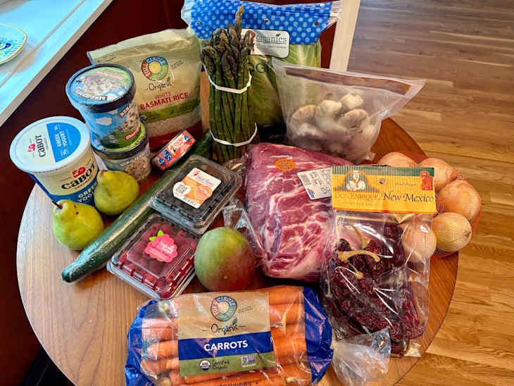 Assorted groceries on a wooden table, including fruits, vegetables, yogurt, rice, and dried peppers.